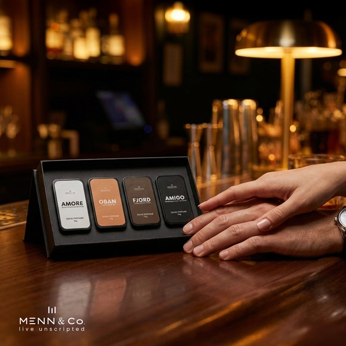 Four cases of Solid Perfume name Amigo, Amore, Oban, Fjord on a stand with 'Menn & Co' branding, placed on a bar counter.