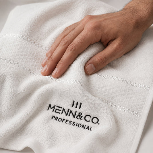 A close-up of a white textured towel with a hand resting on it, featuring the black embroidered logo 'MENN & CO. PROFESSIONAL'.