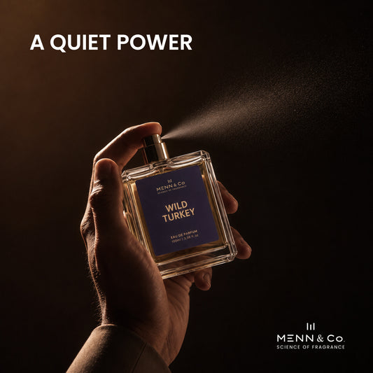 A hand holds a square glass perfume bottle labeled 'MENN & CO. WILD TURKEY' against a dark brown background, with perfume mist spraying from the nozzle.