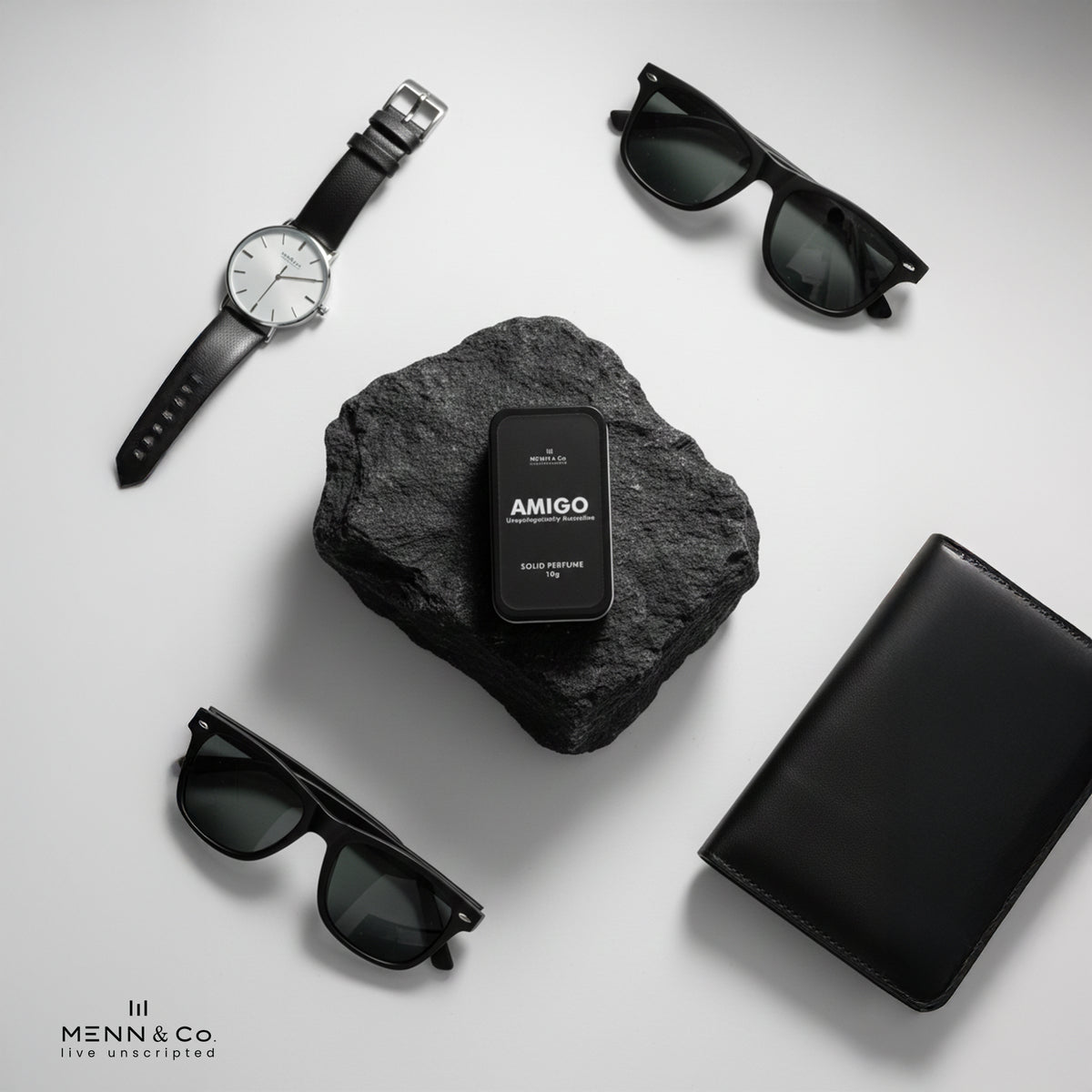 Black watch, sunglasses, and black box with 'AMIGO' on a gray stone surface.