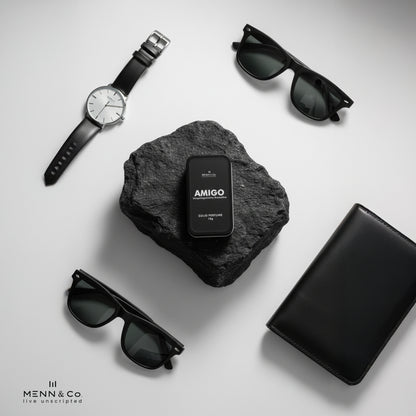 Black watch, sunglasses, and black box with 'AMIGO' on a gray stone surface.
