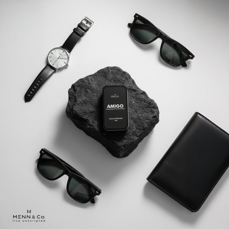 Black watch, sunglasses, and black box with 'AMIGO' on a gray stone surface.