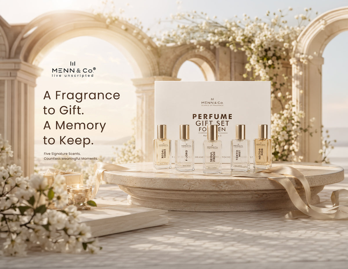 Menn & Co perfume gift set displayed on a stone pedestal in a classical architectural setting with white flowers. The set includes a large white box and five small clear glass perfume bottles with gold caps.