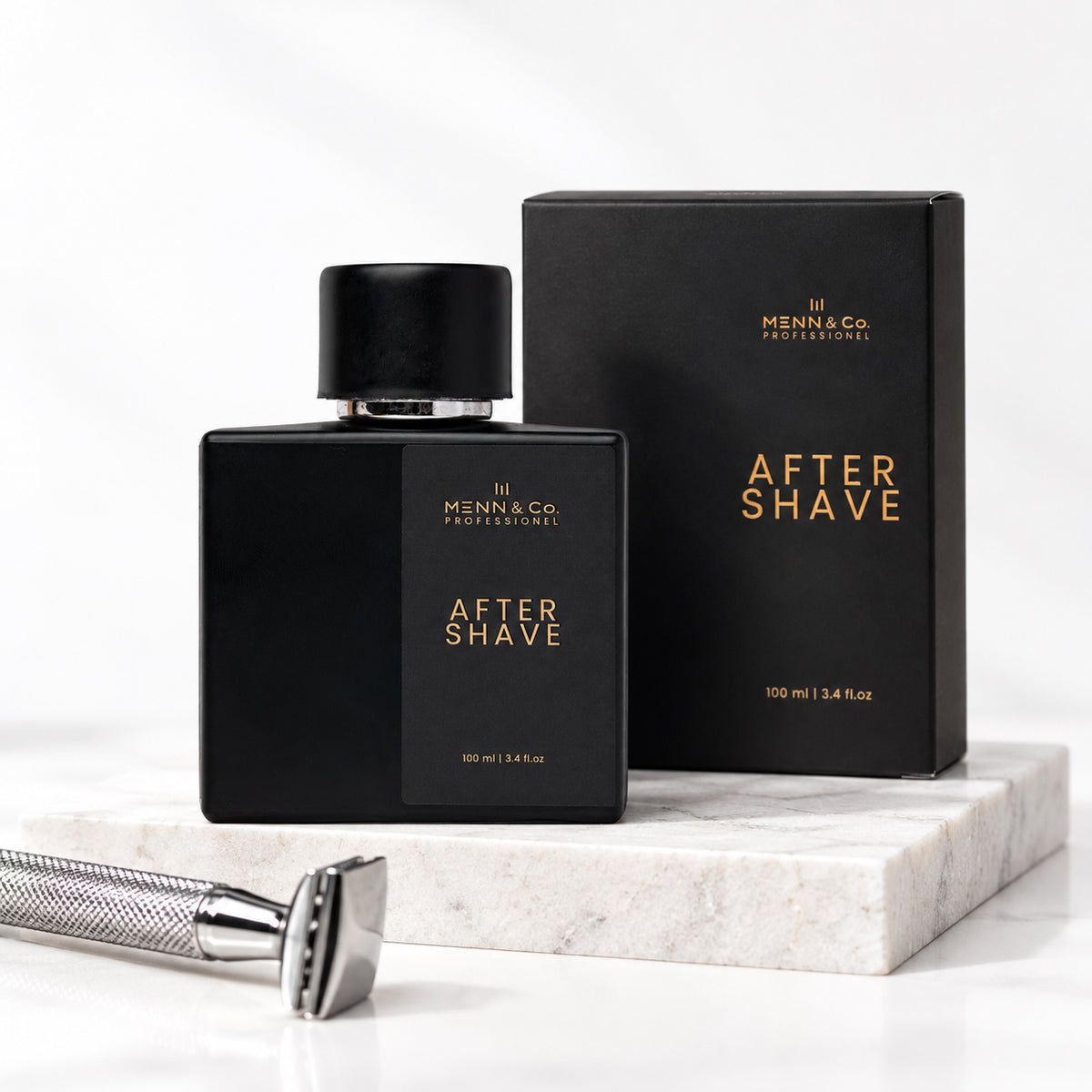 Menn & Co Professional After Shave bottle and box in matte black with gold lettering, displayed on a white marble block with a silver safety razor in the foreground.