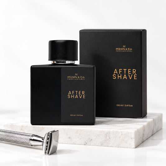 Menn & Co Professional After Shave bottle and box in matte black with gold lettering, displayed on a white marble block with a silver safety razor in the foreground.
