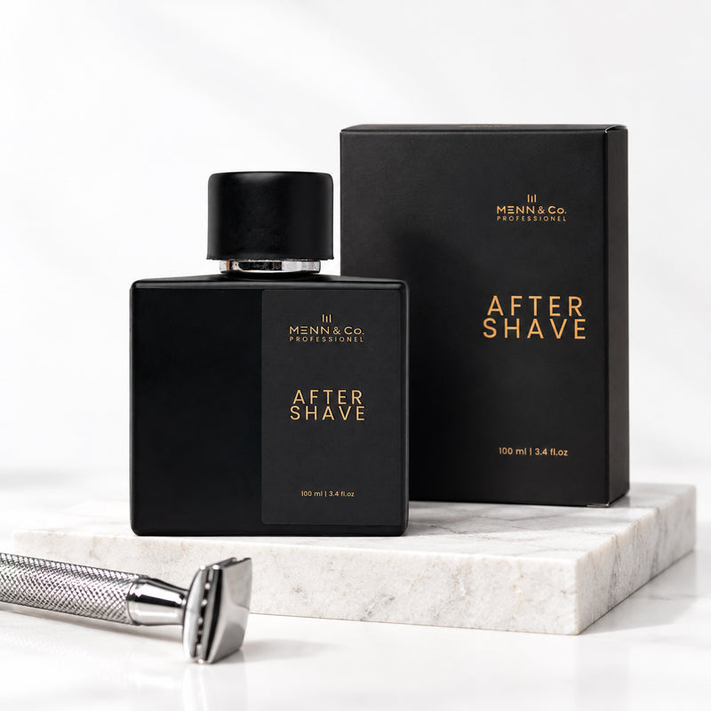 Menn & Co Professional After Shave bottle and box in matte black with gold lettering, displayed on a white marble block with a silver safety razor in the foreground.