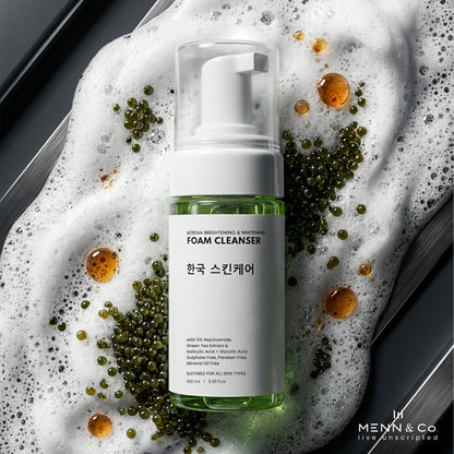 A bottle of Menn & Co. foam cleanser with a white pump and clear cap, filled with green liquid, set on a dark surface surrounded by rich white lather and scattered green tea leaves.
