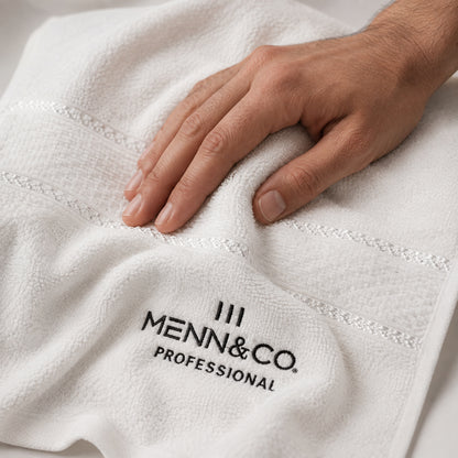 A close-up of a white textured towel with a hand resting on it, featuring the black embroidered logo 'MENN & CO. PROFESSIONAL'.