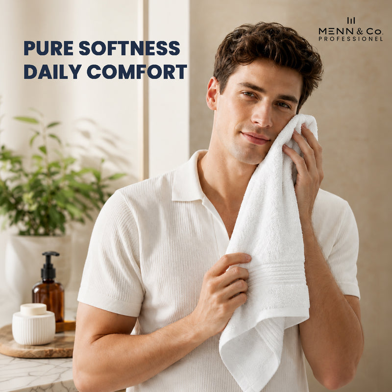 A man in a white ribbed short-sleeve polo shirt holds a white textured towel against his face in a bright bathroom setting. The text 'PURE SOFTNESS DAILY COMFORT' appears on the left, and the 'Menn & Co Professional' logo is in the top right.