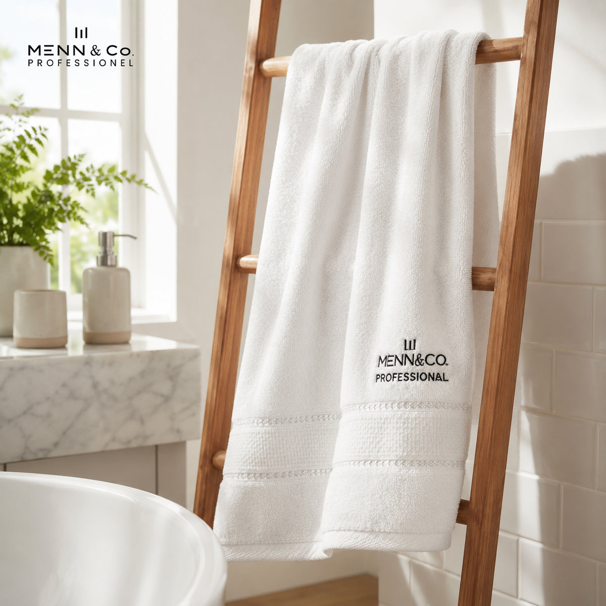 A white plush bath towel with black embroidered 'MENN & CO PROFESSIONAL' branding hangs on a wooden ladder rack in a bright, modern bathroom setting.