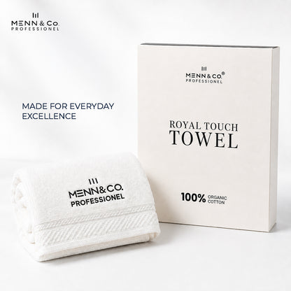 A folded white cotton towel with black printed branding sits in front of its matching off-white product box. The packaging reads 'ROYAL TOUCH TOWEL' and '100% ORGANIC COTTON,' and the towel reads 'MENN&Co PROFESSIONEL.