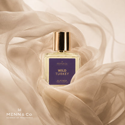 A bottle of Menn & Co Wild Turkey Eau de Parfum centered on draped beige fabric. The bottle is clear glass with pale yellow liquid inside, a metallic gold cap, and a dark purple label with gold lettering.
