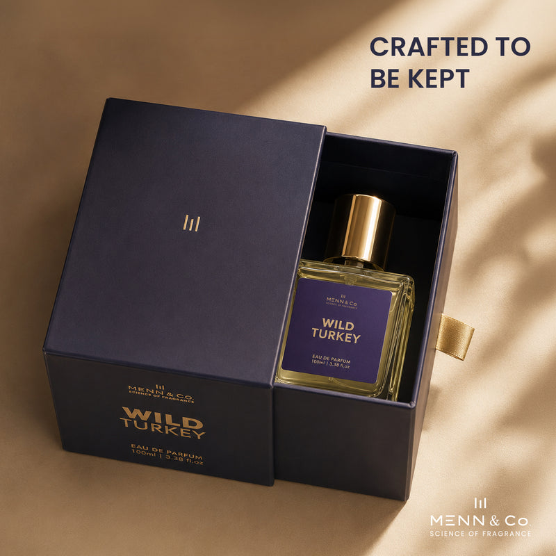 Menn & Co Wild Turkey Eau de Parfum bottle displayed inside its navy blue presentation box, with the lid leaning beside it on a warm beige textured background. The bottle has a gold cap and a purple label, and the box features gold lettering.